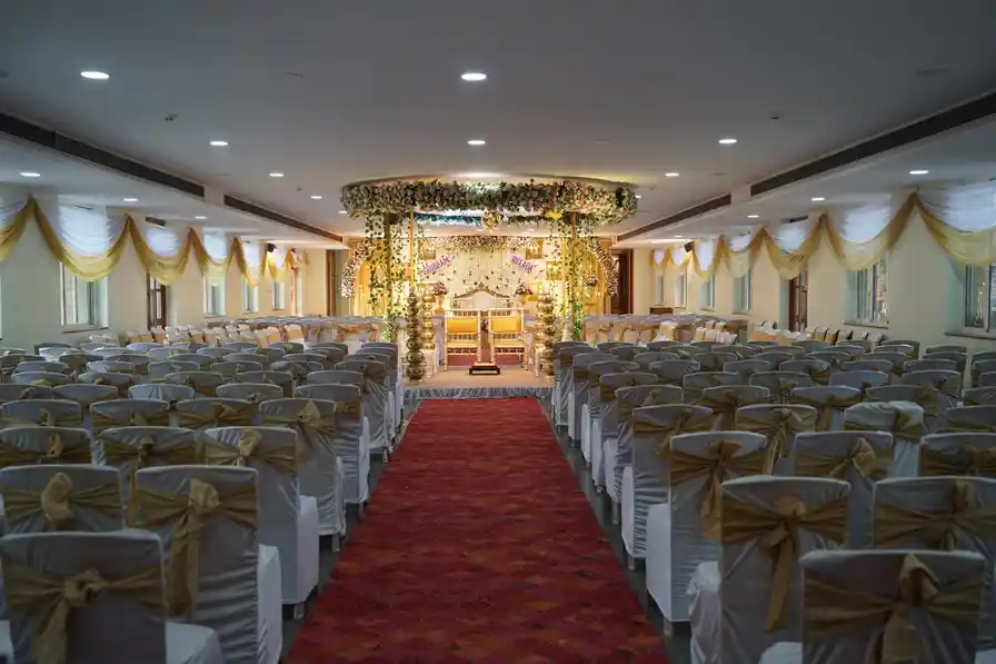 The hall set for a ceremony, a red carpet aisle running between cream-covered chairs to a gold mandap