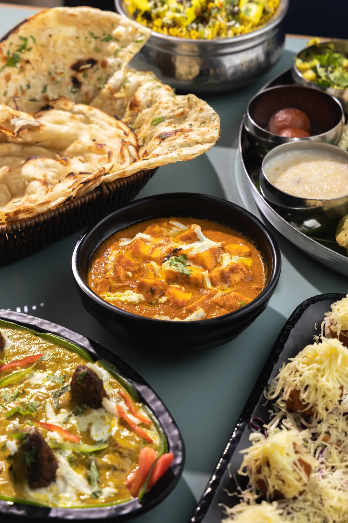 Paneer butter masala in a black bowl, with naan and a thali behind it