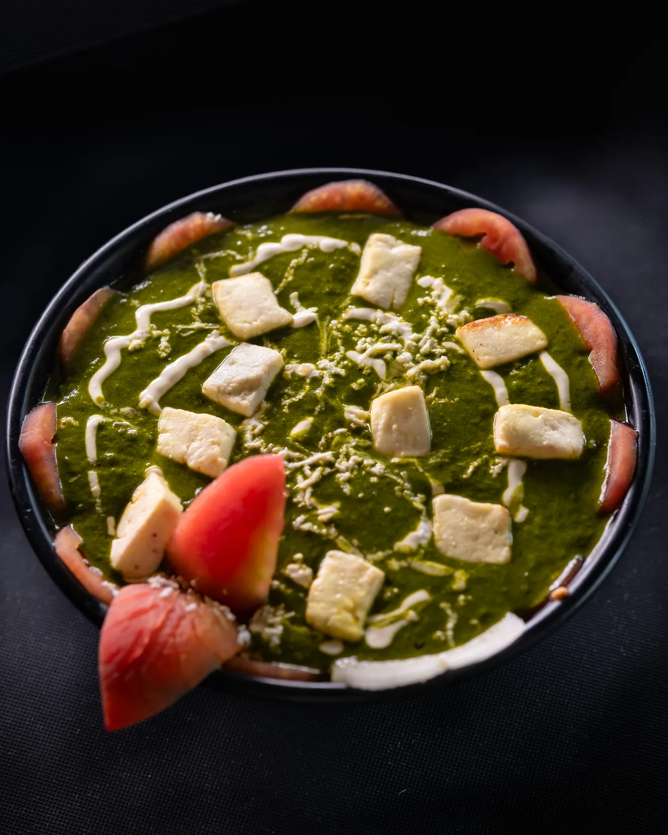 Palak paneer in a round black dish, cream drizzled over the paneer cubes