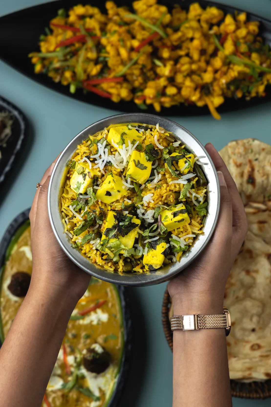 Hands holding a steel bowl of paneer biryani garnished with coconut and coriander